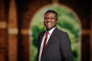 A person in a dark suit and red tie smiles warmly, standing in front of a blurred brick archway with greenery visible through it.